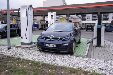 BMW i3 elektrikli araba bir elektrik şarj istasyonundan şarj ediliyor, kablo şarj ediliyor, diğer bir benzin istasyonu arka planda görünen geleneksel benzin arabaları için, Berlin, Almanya - 17 Şubat 2024