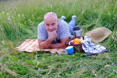 Yetişkin bir adam 60 yaşında, çayır çimenlerde yatıyor, meyveli piknik sepetinin yanında, termoslu çay, aile pikniği konsepti, hayatın tadını çıkar, aktif yaşam tarzı, insan mutluluğu