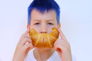 close-up of child's face, boy of 8 years old holds rosy baked croissant with hands near mouth, depicts cheerful smile, concept delicious breakfast, happy childhood, emotional development children