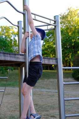 child, boy 8 years old Asian-European appearance with blue hair climbs up on metal sports equipment in playground, concept of sports achievements deniya, striving for success, physical development