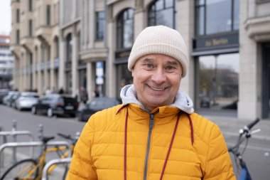 Olgun karizmatik bir adam, 65 yaşında, parlak sarı kabarık ceketli ve bereli, şehir sokaklarında gülümseyen, tarihi binalar arka planda, Avrupa 'da yürüyor.