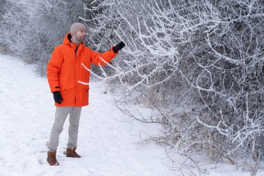 Parlak turuncu ceketli genç adam kışın güzel karlı parkta yürüyor, psikoloji ve etki, mevsimsel mutluluk, açık hava meditasyonu, aktif yaşam tarzı, soğuk hava.