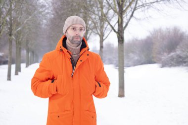 Parlak turuncu ceketli 30 yaşındaki genç adam kışın karlı bir parkta yürüyor, psikoloji ve etki, mevsimsel mutluluk, açık hava meditasyonu, aktif yaşam tarzı, soğuk hava.