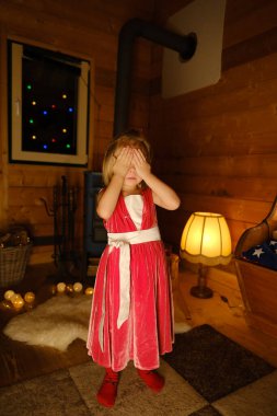 Joyful Child, happy cool girl 4 years old in red dress closing eyes, anticipating gifts by glowing fireplace, Kid eagerly awaiting Christmas surprises on St. Nicholas Day, Christmas coming clipart