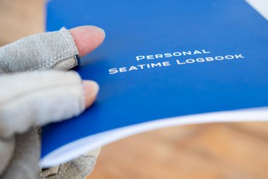 Gloved hand holding blue Personal Seatime Logbook. Maritime record keeping. Seafarer documentation. Nautical career tracking, Seafarer's chronicle, Sea hours documented clipart