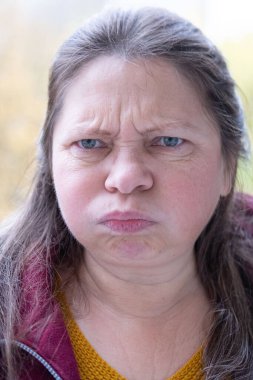 Stresli 50 yaşında bir kadın, çevresi yeşillik ve dingin çevre ile çevrili, negatif duygular sergiliyor, öfke ve gerilimle tepki veriyor, duygusal dengesizlik gösteriyor.