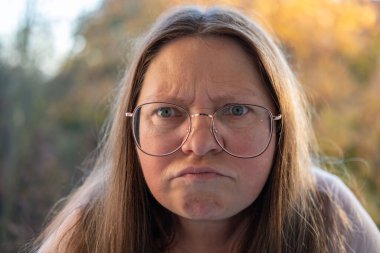 Stresli 50 yaşında bir kadın, çevresi yeşillik ve dingin çevre ile çevrili, negatif duygular sergiliyor, öfke ve gerilimle tepki veriyor, duygusal dengesizlik gösteriyor.