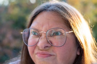 Stresli 50 yaşında bir kadın, çevresi yeşillik ve dingin çevre ile çevrili, negatif duygular sergiliyor, öfke ve gerilimle tepki veriyor, duygusal dengesizlik gösteriyor.