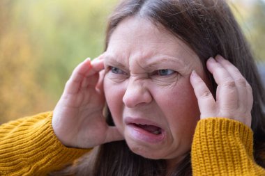 Stresli 50 yaşında bir kadın, çevresi yeşillik ve dingin çevre ile çevrili, negatif duygular sergiliyor, öfke ve gerilimle tepki veriyor, duygusal dengesizlik gösteriyor.