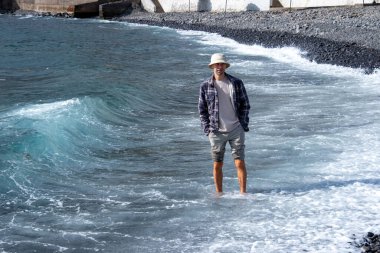 Introspective man stands knee-deep in turquoise waters, Coastal wanderer in checkered shirt and pale bucket hat, experiencing nature's rhythm secluded rocky beach tropical climate zone clipart