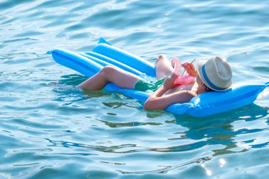 Mutlu çocuk şık bir şapka ve güneş gözlüğüyle güneşleniyor. Lüks plaj gününün tadını çıkarıyor. Genç çocuk şişme salda yüzerken ferahlatıcı bir içecek yudumluyor.