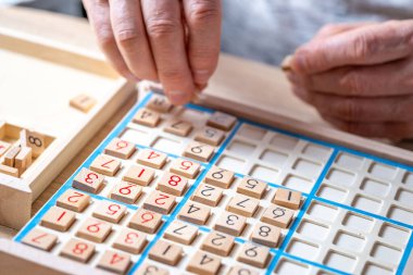 Sayıyı Sudoku yapbozuna dikkatlice yerleştiren eller, meydan okuyucu bulmacaları çözmeye, zihinsel uyarımlara ve klasik oyunlardan keyif almaya kendini kaptırmış yaşlı kişi,