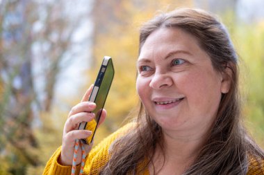 Zarif, 50 yaşında, beyaz bir kadın. Akıllı telefonunu açık hava ortamında kontrol ediyor. Dijital yaş, olgun yaşam tarzı, gerçek insanlar, doğal altyapı.