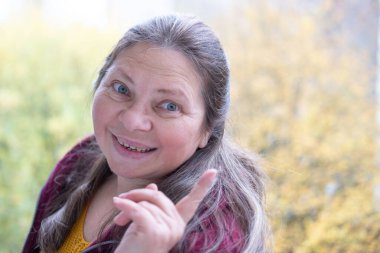 Neşeli, olgun bir kadın eğleniyor, komik bir yüz ifadesi yapıyor, neşeli hareketlerle komik bir resim yapıyor, garip bir ifade sergiliyor, açık havada kendiliğinden çekilen bir an.