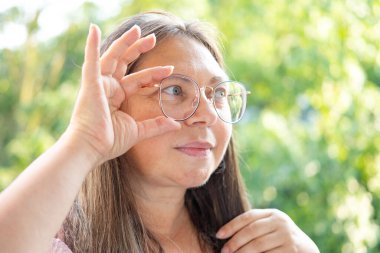 50 yaşında bir kadın, gözlüklerle yan yana bakıyor, görüş muayenesi, göz hastalıklarının tedavisi, yaşlanma ve görme düzeltmesi ihtiyacı.