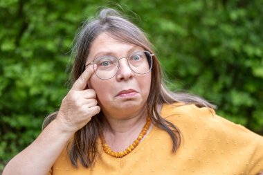 Komik yaşlı kadın alnına dokunuyor, kafası karışmış ve eğleniyor, açık havada, kırışıklıklar, şaşkın yüz, şüpheci bakışlar, yaşlanma karşıtı bakışlar, cilt endişesi, insani duygular, samimi bir portre