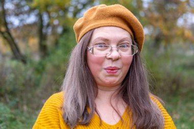 Pozitif orta yaşlı kadın, hardal kıyafetli, temiz havanın tadını çıkarıyor, sonbaharda orman, sonbahar havası, mutluluk doğa yürüyüşü, sonbaharın tadını çıkarıyor, gülümsüyor, neşeli bir ifade, açık havada, açık havada olgun bir portre