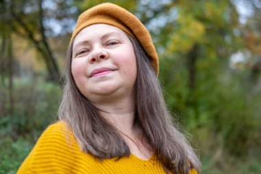 Pozitif orta yaşlı kadın, hardal kıyafetli, temiz havanın tadını çıkarıyor, sonbaharda orman, sonbahar havası, mutluluk doğa yürüyüşü, sonbaharın tadını çıkarıyor, gülümsüyor, neşeli bir ifade, açık havada, açık havada olgun bir portre