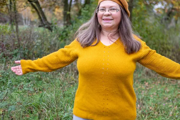 Pozitif orta yaşlı kadın, hardal kıyafetli, temiz havanın tadını çıkarıyor, sonbaharda orman, sonbahar havası, mutluluk doğa yürüyüşü, sonbaharın tadını çıkarıyor, gülümsüyor, neşeli bir ifade, açık havada, açık havada olgun bir portre