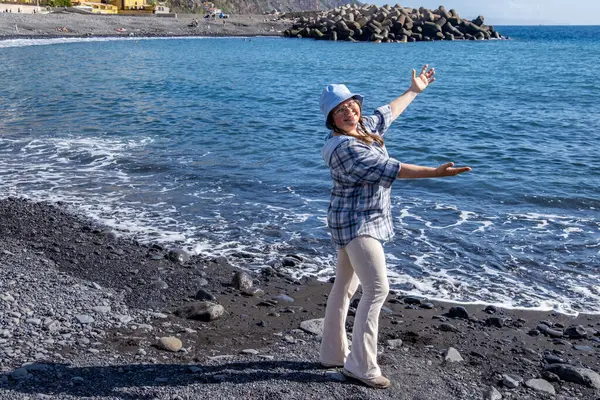 Okyanus kıyısında mutlu, orta yaşlı bir kadın, saçlarında rüzgarı hissediyor, suyun kıyısında zarif yaşlı bir bayan, Madeira adasında Atlantik Okyanusu, sıcak bir gün, doğayla bağlantı, nazik bir gülümseme, sahil yürüyüşü