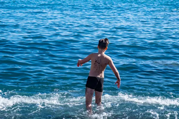 Okyanus dalgalarında oynayan genç çocuk, siyah volkanik kumsalda, denize düşen, neşeli yaz anı, doğal eğlence, sağlıklı çocukluk, yalınayak özgürlük, tropikal doğanın ve güneşin tadını çıkarıyor.