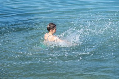 Çocuk denizde neşeyle yüzüyor, yaz tatilinin tadını çıkarırken su güvenliği becerilerini öğreniyor, çocuk deniz dalgalarında oynuyor, kaygısız ve mutlu, ebeveynler güvenliği ve boğulma riskini göze alıyor. 