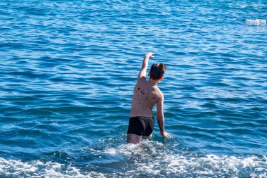 Okyanus dalgalarında oynayan genç çocuk, siyah volkanik kumsalda, denize düşen, neşeli yaz anı, doğal eğlence, sağlıklı çocukluk, yalınayak özgürlük, tropikal doğanın ve güneşin tadını çıkarıyor.