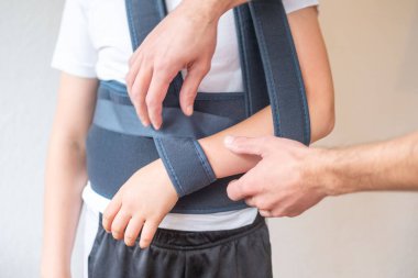 Ortopedik askı takan genç bir çocuk, sağlık hizmeti sağlayıcısının hasta rahatlığı ve iyileşme süreci için ortopedik destek cihazını ayarladığı fiziksel terapi danışmanlığı