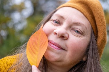 Düşen yapraklı mutlu olgun bir kadın, kadın elinde sarı bir yaprak, kafasında bere, parkta arka plan, orman sakin, zarif yaşlanma, hayat dolu yaşlanma, sağlıklı yaşam tarzı, altın yapraklar, sonbahar havası konsepti