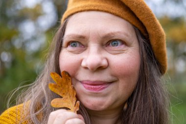 Düşen yapraklı mutlu olgun bir kadın, kadın elinde sarı bir yaprak, kafasında bere, parkta arka plan, orman sakin, zarif yaşlanma, hayat dolu yaşlanma, sağlıklı yaşam tarzı, altın yapraklar, sonbahar havası konsepti