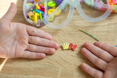çocuk renkli boncuklarla oynuyor, çocuğun elleri dikkatle ipe renkli boncuklar takıyor, boncuklama aktivitesi, güzel motor beceriler geliştiriyor, çocukluk dönemi yaratıcılığı, eşsiz mücevherler yaratıyor.