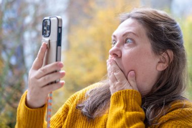 Şok olmuş, şaşırmış olgun bir kadın akıllı telefonunu tutar ve geniş gözleriyle duygusal tepki verir, şok içinde ekrana bakar. Beklenmedik haberlere duygusal tepki, virüs içeriği, beklenmedik online olay