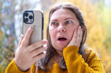 Şok olmuş, şaşırmış olgun bir kadın akıllı telefonunu tutar ve geniş gözleriyle duygusal tepki verir, şok içinde ekrana bakar. Beklenmedik haberlere duygusal tepki, virüs içeriği, beklenmedik online olay