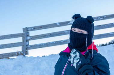 Kış kıyafetleri içinde, siyah örgü şapkalı, ponpon ve yüz maskeli neşeli genç bir kadın, karlı bir günde kırmızı kızak tutarken gülümsüyor, kış neşesi, açık hava macerası, soğuk hava modası