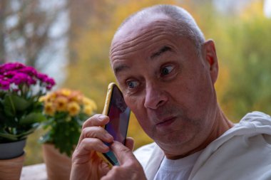 Şok olmuş, şaşırmış, olgun bir adam akıllı telefonunu tutuyor ve geniş gözleriyle duygusal tepkiler vererek ekrandaki şoka bakıyor. Beklenmedik haberlere duygusal tepki, virüs içeriği, beklenmedik online olay