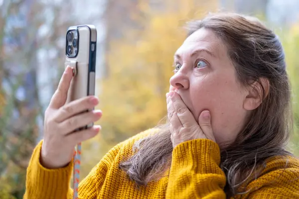 Şok olmuş, şaşırmış olgun bir kadın akıllı telefonunu tutar ve geniş gözleriyle duygusal tepki verir, şok içinde ekrana bakar. Beklenmedik haberlere duygusal tepki, virüs içeriği, beklenmedik online olay