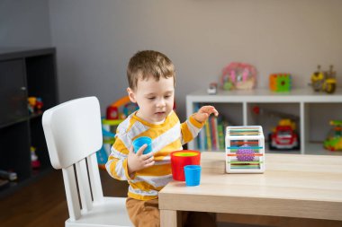 İki yaşında küçük tatlı bir çocuk çocuk çocuk masasında oturur ve odasında eğitici oyuncaklarla oynar. Duyusal renkli toplar ve çok renkli piramitlerin ayrıntıları. Yumuşak seçici odak