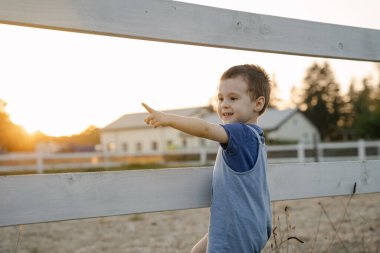 İki ya da üç yaşındaki mutlu çocuk güneşli bir yaz gününde çitin yanında durup parmağıyla işaret ediyor. Yaz gün batımı ışığı. At çiftliği.