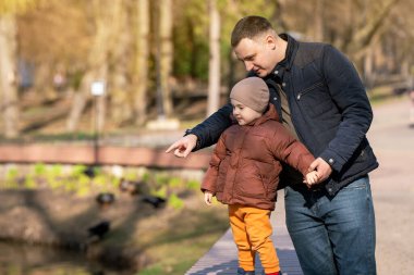 Baba ve küçük çocuk güneşli bir bahar gününde şehrin göl kenarındaki bir parkta dikiliyorlar. Aile hayatı konsepti. Zaman harcamak