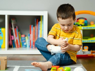 Küçük bir çocuk elleriyle bacağını alçıda tutuyor. Çocuklarda ayak ve parmak kırığı. İnsan sağlığı ve tıp konsepti. Bandaj