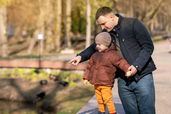 Father and toddler son stand in a park near the city lake on a sunny spring day. Family time lifestyle concept. Spending time