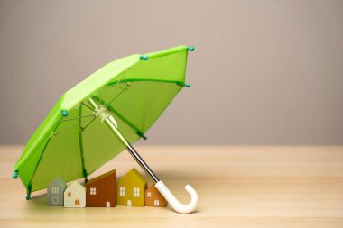 Emlak ve ev sigortası konsepti. Ahşap evler yeşil bir şemsiyenin altında duruyor. Finansal güvenlik. Kapsamlı bir kayıp durumunda onarımlar veya yeniden inşa.
