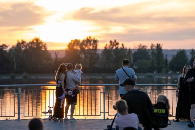 Genç bir aile, bir yaz günü akşam saatlerinde gölün kenarında selfie çekiyor. Akşam kenti boyunca yürü. Yaz zamanı. Ivano-Frankivsk, Ukrayna - 4 Ağustos 2024.