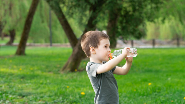 Three year old caucasian toddler boy drinks water from a bottle in the park on a spring or summer day.