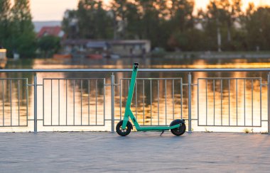 Güneşli bir günde yeşil elektrikli scooter gölün yakınındaki bir yaz parkına park edilir. Şehirde dolaşıyordu. Turizm ve seyahat.