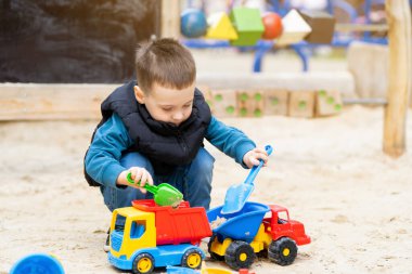 Küçük tatlı çocuk üç yaşında bir bahar günü kum havuzunda oynuyor. Çocuklar için açık hava gelişim aktiviteleri. Oyuncak arabalar ve kürek.