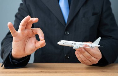 Bir adam elinde bir uçak tutuyor ve tamam işaretini gösteriyor. Pozitif seyahat deneyimleri. Uçuş güvenliği, uygun fiyatlı uçak biletleri, pürüzsüz hava yolculuğu, tatil planlama ve havayolu promosyonları..