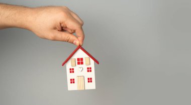 Gri arka planda kırmızı çatılı bir evi tutan bir el. Gayrimenkul, uygun fiyatlı konut kavramı. Konut binalarının değeri. Emlakçı servisi. Satın alma ve satma. İpotek ve kredi