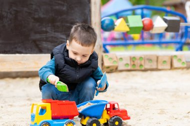 Küçük tatlı çocuk üç yaşında bir bahar günü kum havuzunda oynuyor. Çocuklar için açık hava gelişim aktiviteleri. Oyuncak arabalar ve kürek.