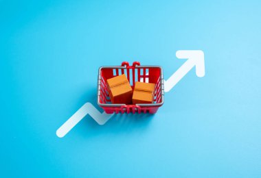 Kutulu mallarla dolu bir alışveriş arabası ve yukarı bakan bir ok. Tüketim artışı. Yüksek fiyatlar, yükselen enflasyon. Satın alma davranış trendleri. Yaşam maliyeti ve piyasa analizi.